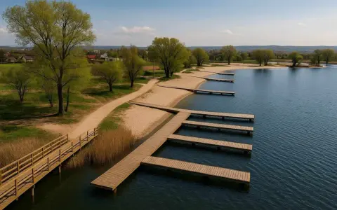 Drewniane pomosty w kształcie litery L i proste, prowadzące do wody, na piaszczystej plaży. W tle zielone drzewa i zabudowania. Idealne miejsce na wypoczynek nad wodą, jak na przylasku ruskim.