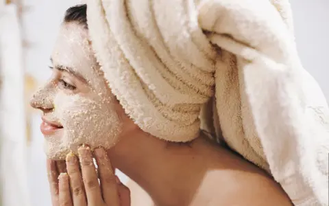Frau mit Handtuch auf dem Kopf trägt Gesichtsmaske und massiert sie sanft ein.