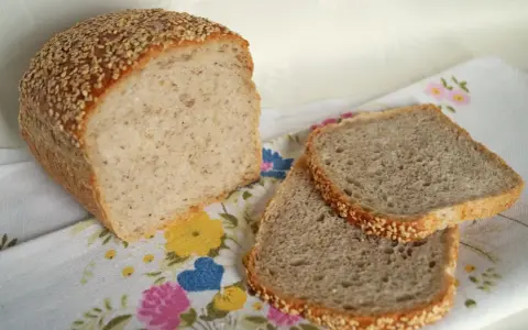 Chleb z kaszą gryczaną, posypany sezamem, leży obok dwóch kromek na kwiecistym obrusie.
