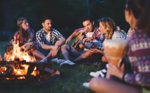 Przyjemne zabawy przy ognisku. Grupa przyjaciół siedzi na trawie, jeden gra na gitarze, inni słuchają.