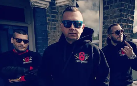 Trzech mężczyzn w bluzach z logo "Gang Albanii". Jeden z nich pokazuje gest ręką.