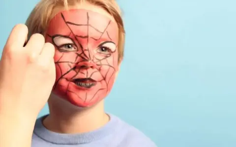 Ein Kind wird mit einem Spiderman-Gesicht bemalt. Rote Farbe und schwarze Spinnennetzmuster verwandeln das Gesicht in eine Superheldenmaske.