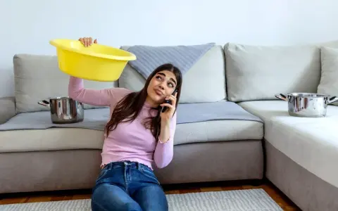 Mujer preocupada llama por teléfono mientras sostiene un barreño amarillo para recoger agua de las goteras en casa. Ollas esperan en el sofá.