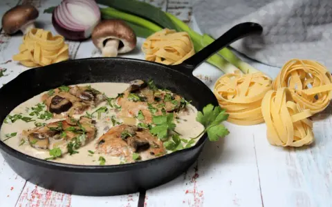 Ein köstliches Schnitzelpfanne Rezept mit cremiger Pilzsoße in einer Gusseisenpfanne, garniert mit Petersilie. Daneben liegen frische Pasta-Nester, Pilze und Lauch.