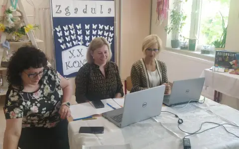 Trzy kobiety pracują przy stołach w sali, która wygląda jak szkoła specjalna. Na ścianie wisi tablica z motylami i napisem "Zgadula".