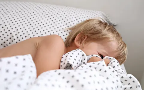 Niño rubio se esconde bajo la sábana. Aprende como hacer que un niño duerma solo con paciencia y rutinas.