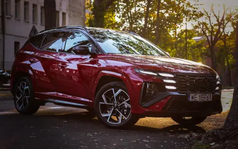 Czerwony Hyundai Tucson Hybrid zaparkowany na tle jesiennych drzew i kamienicy.