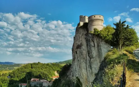 La fortezza di San Leo domina un picco roccioso, con un borgo sottostante e un cielo azzurro punteggiato di nuvole.