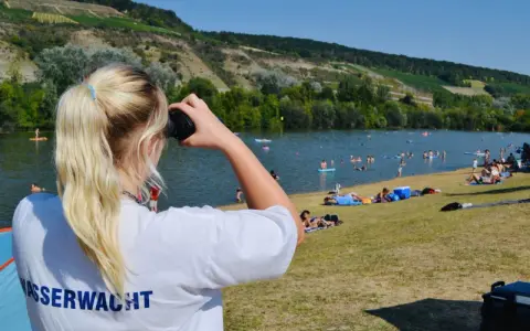 Wasserwacht am Erlabrunner Badesee: Eine junge Frau mit blondem Pferdeschwanz blickt durch ein Fernglas über den belebten See, auf dem viele Menschen schwimmen und sonnen.