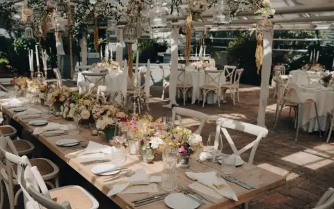 Romantische Hochzeitslocation Ideen: Ein langer Holztisch mit Blumenarrangements, Kerzen und weißen Stühlen, bereit für Gäste.