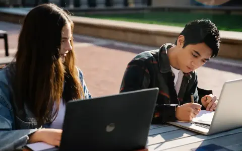Dwoje studentów korzysta z laptopów na zewnątrz, przygotowując się do zajęć. To idealny przykład elastyczności, jaką oferują studia hybrydowe.