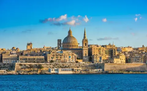 Malta, wyspa do kogo należy? Widok na Vallettę z kopułą i wieżą kościelną, otoczoną zabytkową architekturą, nad błękitnym morzem.