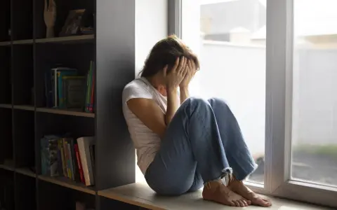 Kobieta siedzi przy oknie, zakrywając twarz dłońmi. Wokół panuje nastrój melancholii, odzwierciedlający dystymię.