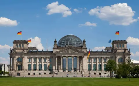 Imponujący Reichstag z charakterystyczną szklaną kopułą, otoczony zielonym trawnikiem i błękitnym niebem z chmurami.