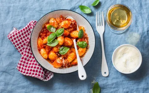 Pyszne kluski gnocchi w sosie pomidorowym, udekorowane bazylią. Obok kieliszek białego wina i miseczka parmezanu.