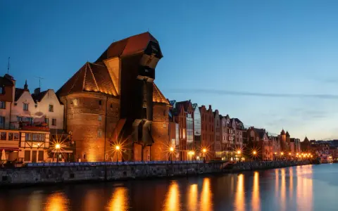Gdańsk nocą. Historyczny Żuraw i kamienice nad Motławą, rozświetlone latarniami, odbijają się w wodzie.