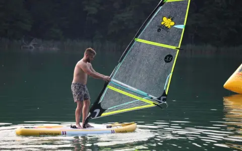 Mężczyzna na desce z żaglem, gotowy do wind sup. Na żaglu pszczółka i napis "3.5".