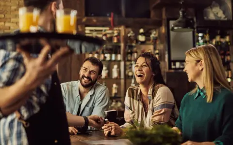 Un camarero sirve bebidas a un grupo de amigos riendo en un bar. La conversación animada sugiere que aún no es tarde para el ruido.