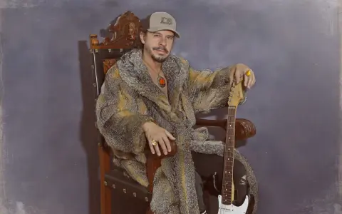 Luis Brea, con gorra y abrigo de piel, sentado en un trono con una guitarra.