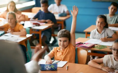 Chłopiec z podniesioną ręką czeka na ocenianie odpowiedzi. Inne dzieci pracują przy ławkach.
