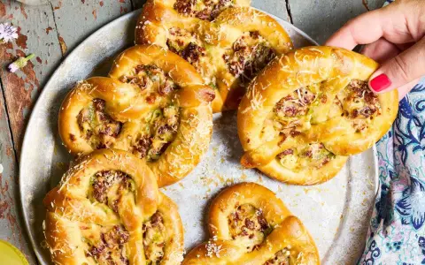 Herzhafte Brezeln mit Lauch und Speck, die an Flammkuchen erinnern, liegen auf einem Blech. Eine Hand greift nach einer Brezel.