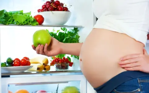 Gravid kvinna tar ett grönt äpple ur kylen fylld med färsk frukt och grönsaker. Hon njuter av hälsosam mat under sin soja graviditet.