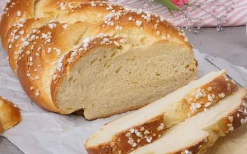 Ein goldbrauner Hefezopf, bestreut mit Zuckerkristallen, liegt auf Backpapier. Ein Stück ist angeschnitten, die weiche Krume ist sichtbar. Ein tolles Rezept für Hefezopf!