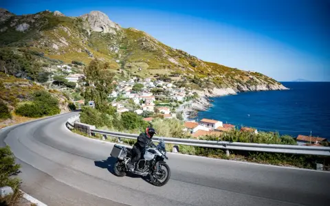 Un motociclista esplora le curve panoramiche dell'Elba, con il mare blu e un villaggio pittoresco sullo sfondo.