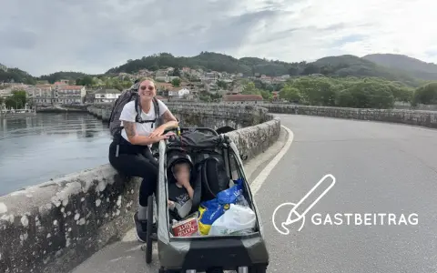 Frau mit Rucksack und Kinderwagen auf einer Brücke, bereit für eine abc tour. Im Hintergrund eine Stadt und Berge.