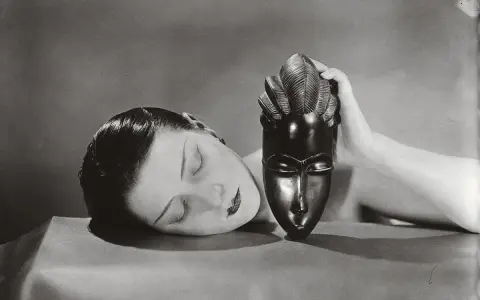 Foto Man Ray: volto femminile addormentato accanto a una maschera africana tenuta da una mano.