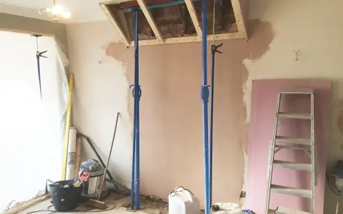 Travaux de rénovation : des étais soutiennent une structure en bois au-dessus d'une zone où l'on s'apprête à casser une cheminée. Matériaux de construction et outils au sol.