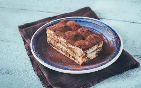 Pyszny kawałek prawdziwego tiramisu, posypany kakao, na niebieskim talerzu.