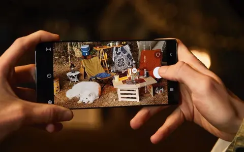 Une photo de téléphone capture un chien blanc endormi lors d'un camping. L'écran du téléphone montre une scène de camping avec une tente, une chaise pliante et un feu de camp.