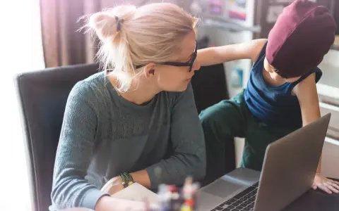 Une mère travaille sur son ordinateur portable, son enfant curieux s'approche. Un moment de vie quotidienne illustrant l'équilibre digital.