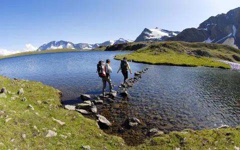 Due escursionisti attraversano il lago di Ceresole su un sentiero di pietre, con montagne innevate sullo sfondo.