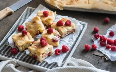 Słodkie brownie i blondie z malinami, pokrojone na kwadraty, ułożone na tacy. Kilka malin rozsypanych obok.