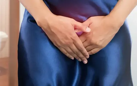 Kobieta w granatowej sukience z rękami na podbrzuszu, w tle toaleta. Problem z nietrzymaniem moczu? Czas na wizytę u lekarza.