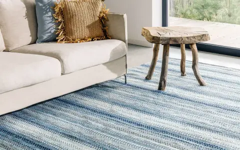 En ljus soffa med kuddar och ett rustikt sidobord står på en randig matta i blåa nyanser. Olika typer av mattor kan skapa en mysig atmosfär.