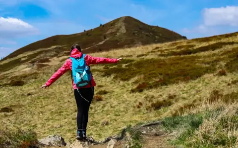 Kobieta w różowej kurtce i turkusowym plecaku na szlaku w Bieszczadach. Przed nią wznosi się góra.