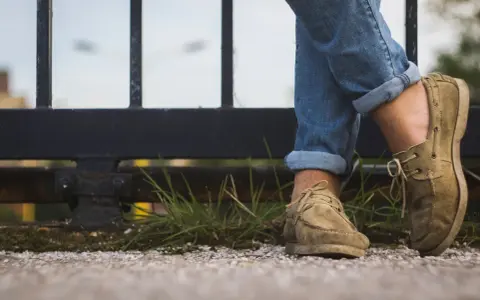 Gambe con jeans arrotolati e scarpe senza calzini, appoggiate su un muretto con erba.