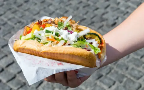 Kebab w bułce z mięsem, warzywami i serem feta, trzymany w dłoni.
