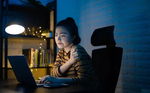 Une jeune femme, en pleine **mise en place télétravail**, ressent une douleur à l'épaule devant son ordinateur, éclairée par une lampe de bureau.