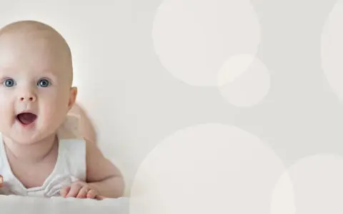 Uśmiechnięte niemowlę 4 miesiące leży na brzuchu, z otwartymi ustami i szeroko otwartymi oczami, patrząc wprost na widza.