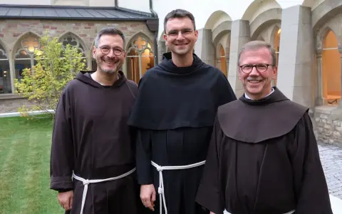Drei Franziskaner Minoriten in braunen und schwarzen Kutten lächeln in die Kamera vor einer Klosterfassade.
