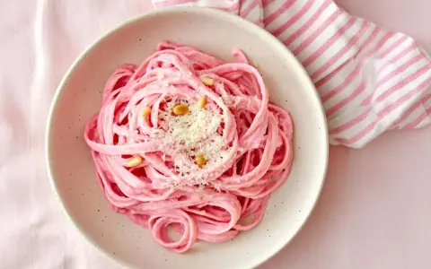 En skål med rosa pasta, toppad med pinjenötter och riven ost. En randig duk i rosa och vitt ligger bredvid.