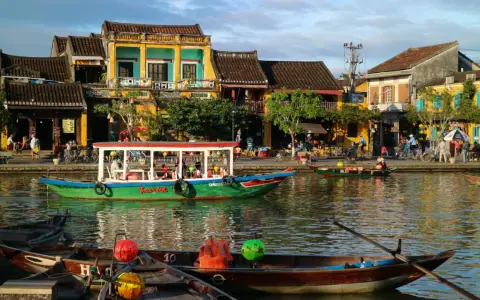 Båtar flyter på floden i Hoi An, Vietnam, med färgglada hus och folk som promenerar längs kajen.