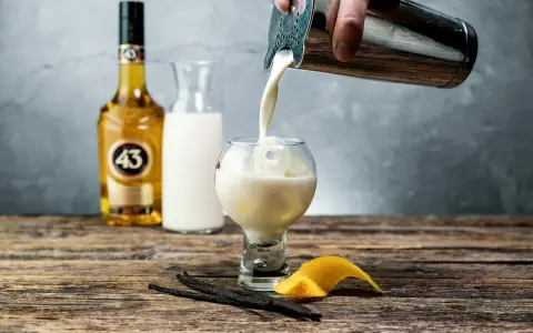 En hand häller en krämig dryck från en shaker i ett glas med is. Bredvid står en flaska Licor 43, en karaff med mjölk, vaniljstänger och citronskal.