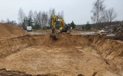 Żółty koparka wykonuje wykopy pod fundament. W tle widać stosy piasku i drzewa.