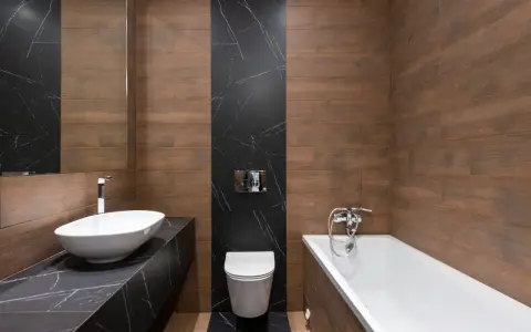 Baño moderno con lavabo ovalado, bañera y un inodoro con salida vertical o horizontal, todo rodeado de azulejos de madera y mármol negro.