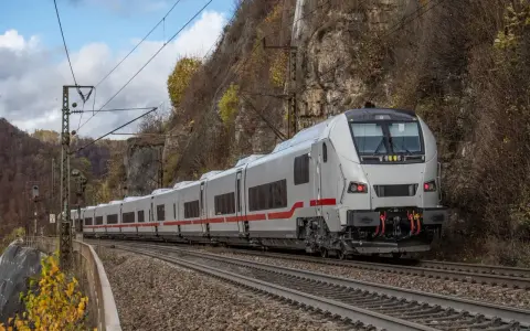 Ein weißer ICE-Zug mit roten Streifen fährt auf Gleisen an einer Felswand entlang. Herbstlaub färbt die Szenerie.